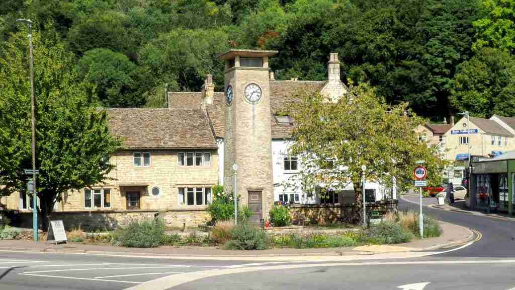 Nailsworth war‑memorial clock tower in a small garden. Grade II–listed Nailsworth war‑memorial clock tower with four‑faced clock and plaques set in a small floral garden at a road junction, built in 1952 to replace an earlier memorial.